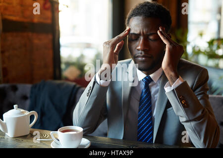 Ritratto di bello americano africano l uomo che soffre di mal di testa e affaticamento al tavolo nel ristorante moderno durante la pausa caffè Foto Stock