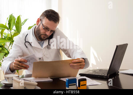 Un dottore seduto alla sua scrivania. Parlando al telefono da rivedere la cartella clinica del paziente Foto Stock