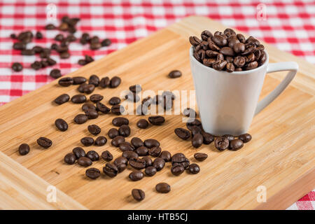 Un bianco tazza di caffè con i chicchi di caffè in legno e un colore rosso-bianco tovaglia a scacchi in background. Foto Stock