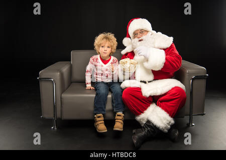 Babbo Natale mangiare popcorn con il bambino e seduto sul divano su nero Foto Stock