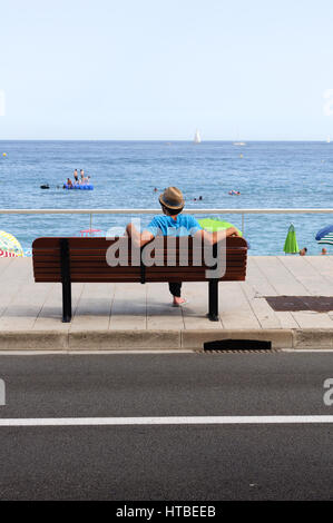 Vista posteriore di un uomo con un cappello su una panchina guardando la spiaggia di Menton, Francia Foto Stock