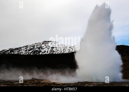 Strokkur Geyser, chiamato anche il grande Geysir, in Islanda in eruzione di fronte ad una montagna innevata Foto Stock