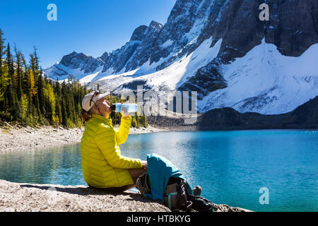 Escursionista femmina bere da una bottiglia d'acqua sui rocciosi lago alpino litorale con nevicato scogliere di montagna in background Foto Stock