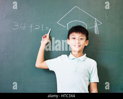 Asian scuola elementare ragazzo che sta sotto un cappello di dottorato disegnato con un gessetto sulla lavagna. Foto Stock