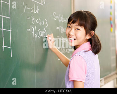 Asian schoolgirl elementari guardando fotocamera a sorridere mentre la soluzione di un problema di matematica. Foto Stock