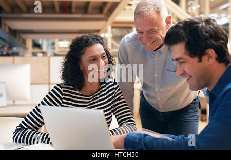 Gruppo diversificato di casualmente vestito sorridente colleghi professionisti parlare insieme su un computer portatile mentre avente un incontro a una scrivania in un ufficio moderno Foto Stock