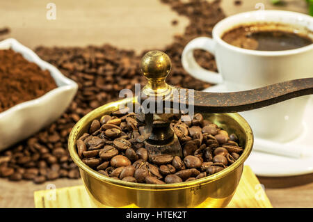 Macinino da caffè con caffè in grani, una tazza di caffè e caffè macinato nella ciotola sul tavolo di legno Foto Stock