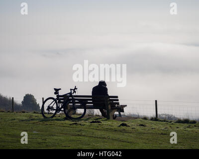 una silhouette di ciclista solitaria agganciata seduta da sola sulla panca Cima della collina Downs che guarda verso la nebbia nebbia nella valle sottostante Foto Stock