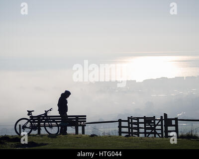 profilo di una silhouette di un elmetto da ciclista solitario in piedi da solo sulla parte superiore Di Downs collina con stile guardando verso la nebbia nebbia nella valle e mare sottostante Foto Stock