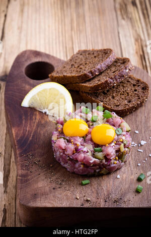 Tartare di manzo sagomato in stampo ad anello, guarnita con due uova di quaglia tuorli e servita con pane di segale sul tagliere di legno Foto Stock