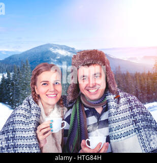 Amore giovane avvolto in caldi plaid con tazze di bevanda calda in mani contro le montagne innevate e la foresta di pini su sfondo Foto Stock