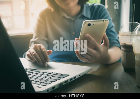 Donna holdong mano smartphone mentre si lavora con il computer portatile in una caffetteria. Lavoratore Freelance sulla giornata lavorativa in linea con la concezione di business con vintage Foto Stock