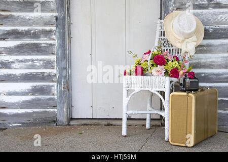 Vecchia macchina fotografica e la valigia con cesto fiorito e il cappello sulla sedia di vimini dalla porta di legno Foto Stock