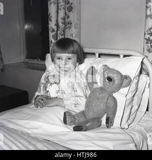 Degli anni Cinquanta, storico, piccola ragazza seduta fino in una cornice di metallo letto di ospedale con un sorriso raggiante, tenendo la sua bambolina e con il suo orsacchiotto di peluche accanto a lei, Inghilterra, Regno Unito. Foto Stock