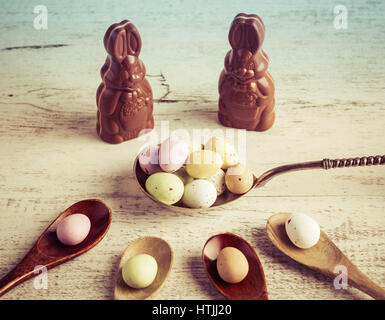 Pasqua rivestite di zucchero candito uova su legno e argento cucchiai con coniglietti di cioccolato bianco su sfondo di legno Foto Stock