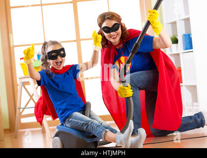 Il bambino e la madre vestita come supereroi utilizzando aspirapolvere in camera. Famiglia di mezza età della donna e la figlia hanno un divertimento durante la pulizia del pavimento. Foto Stock