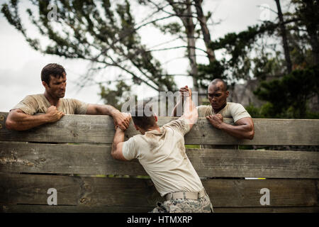 Soldati aiutare l uomo a salire la parete in legno in boot camp Foto Stock