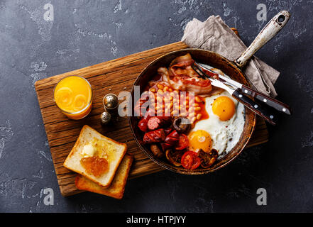 Prima colazione inglese nel recipiente di cottura con uova fritte, salsicce, pancetta, fagioli, pane tostato e succo di arancio scuro su sfondo di pietra Foto Stock