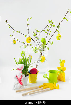 Composizione di pasqua del colore del suo appesi ai rami di betulla con foglie, due conigli e cura per il giardino Foto Stock