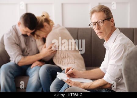 Vero professionista. Grave fiducioso terapeuta maschio guardando a voi e prendere appunti mentre si lavora nel suo ufficio Foto Stock