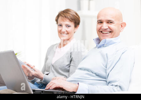 Felice di mezza età matura in un momento di relax a casa con i loro dispositivi elettronici seduti fianco a fianco su un divano con la messa a fuoco per l'uomo sorridente Foto Stock