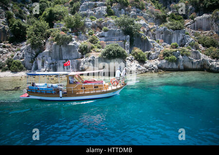 Imbarcazione turistica passando dalla città sommersa di Kekova sotto bandiera turca vicino a rocce Foto Stock