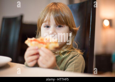 Tre anni bionda bambino affamato di mangiare la pizza e che mostra un pezzo con la sua mano, seduta in marrone scuro sedia indoor Foto Stock