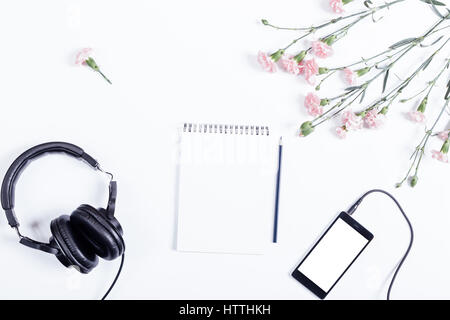 Smart phone, cuffie, blocco di carta, matita e fiori giacciono su un tavolo bianco, vista dall'alto Foto Stock