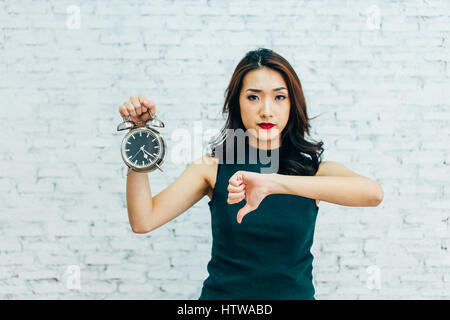 Asian business donna che mostra il pollice verso il basso e tenendo premuto alarm clock - che indica che non è soddisfatto con la gestione del tempo Foto Stock