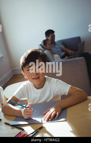 Ritratto di felice ragazzo disegno in carta mentre i genitori usando il computer portatile in background Foto Stock