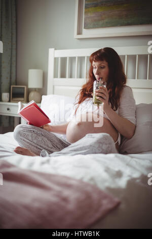 Donna incinta di bere succo di frutta durante la lettura di un libro sul letto Foto Stock