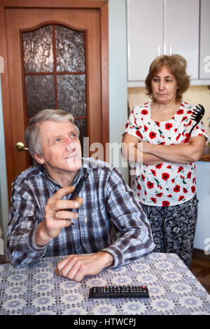 Senior uomo guarda con il telecomando del televisore mentre è seduto al tavolo della cucina, severa moglie sta dietro a bracci incrociati Foto Stock