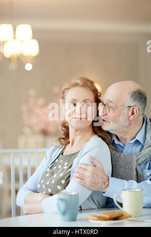 Ritratto di sorridente coppia matura home al tavolo da cucina: uomo anziano abbracciando donna elegante tiepido e sussurra complimenti al suo orecchio Foto Stock
