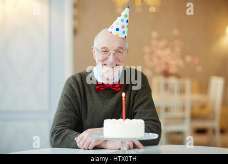 Ritratto di felice vecchio uomo sorridente alla fotocamera mentre è seduto a tavola con la torta di compleanno e indossando party hat Foto Stock