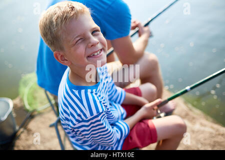 Ritratto di Allegro ragazzino, indossa una camicia a righe, strizzare gli occhi al sole e sorridente guardando la fotocamera mentre la pesca con il padre sul lago Foto Stock