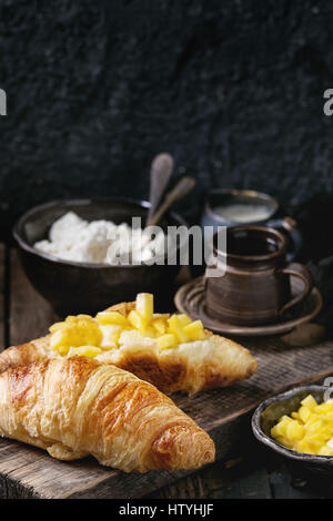 La prima colazione con due cornetti, burro, formaggio, caffè e fette di mango fruit, servita su legno tagliere sopra il vecchio sfondo di legno. Chiudere u Foto Stock