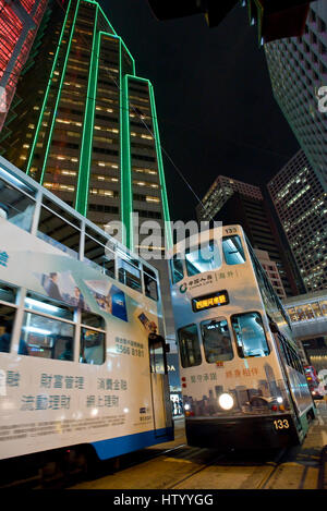 Una vista astratta di 2 Hong Kong tram incontro percorrendo una strada nel quartiere degli affari con i grattacieli sullo sfondo di notte. Foto Stock