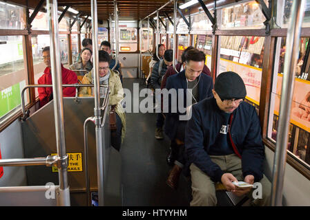 Passeggeri controllare i loro telefoni cellulari seduti all'interno di un tram che viaggia lungo una strada a Causeway Bay, Isola di Hong Kong di notte con Foto Stock