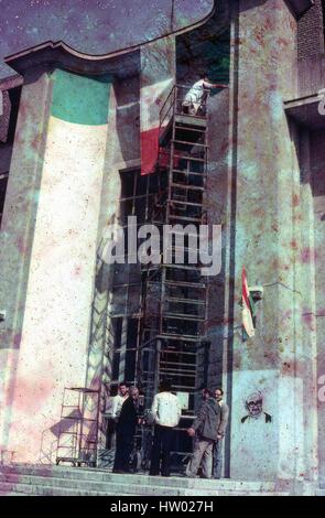 Vista di un uomo pittura i colori nazionali dell'Iran sul lato di un edificio, marzo 1983. Al di sotto di lui un gruppo di uomini in piedi accanto al ponteggio converse accanto ad uno spray dipinto graffiti immagine della rivoluzione iraniana leader Montazeri Hussein-Ali. Danni estesi su foto originale. Foto Stock