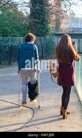 Uomo con cattiva gamba camminando sulle stampelle mentre la ragazza si ferma a prendere una foto a Longleat Safari & Adventure Park, Wiltshire, Regno Unito nel mese di dicembre Foto Stock