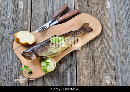 Un affumicato trota arcobaleno servita su un tagliere di legno con rafano, prezzemolo, limone e baguette Foto Stock