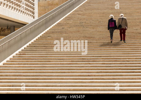 Due vecchie persone che camminano mano nella mano insieme i gradini di pietra di Renzo Piano progettato di entrata e il palazzo del parlamento presso la città di La Valletta Foto Stock