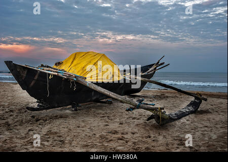 India, Goa. La mattina presto sulla spiaggia Colva. Rising Sun si illumina le nuvole. Barca da pesca, tirando fuori sulla sabbia. Foto Stock