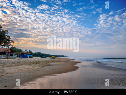 India, Goa. La mattina presto sulla spiaggia Colva. Rising Sun si illumina le nuvole. Barca da pesca, tirando fuori sulla sabbia. Foto Stock
