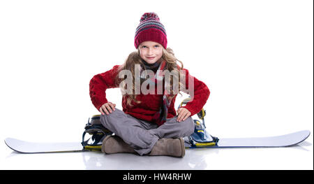 Sorridente bambina con capelli ricci indossare pullover a maglia, sciarpa e cappello seduto su blu snowboard, isolato su bianco. Abbigliamento invernale e sport co Foto Stock