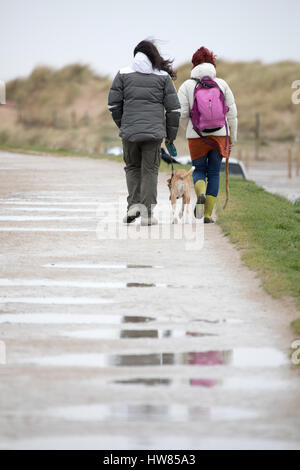 Bagnato e condizioni di vento con due cane femmina gli escursionisti a piedi verso talacre spiaggia con il loro cane al guinzaglio, Flinthsire, Wales, Regno Unito Foto Stock