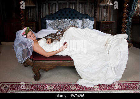 Giovane donna che indossa un bianco abito da sposa, sdraiata su un divano, guardando una rosa rossa in mano Foto Stock