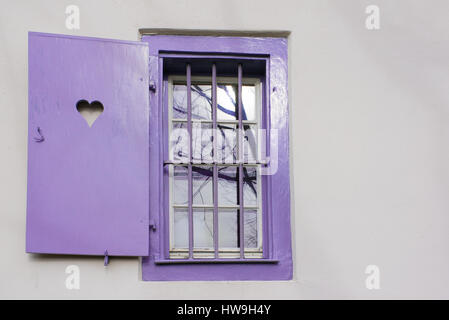 Luce pastello viola finestra nel telaio di legno a forma di cuore sulla serranda e reticolo di ferro su una parete bianca Foto Stock
