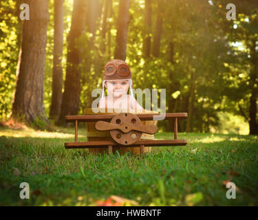 Un piccolo bambino è seduto in un aereo di legno cesto prop nel parco fingendo di viaggiare e di volare con un pilota hat su per una creatività o immaginazione Foto Stock