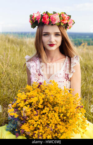 Bella moda ragazza attraente con grande ghirlanda di bellissimi fiori luminosi su i suoi capelli, testa. Seduta in campo ragazza carina con grande mazzo ful o Foto Stock